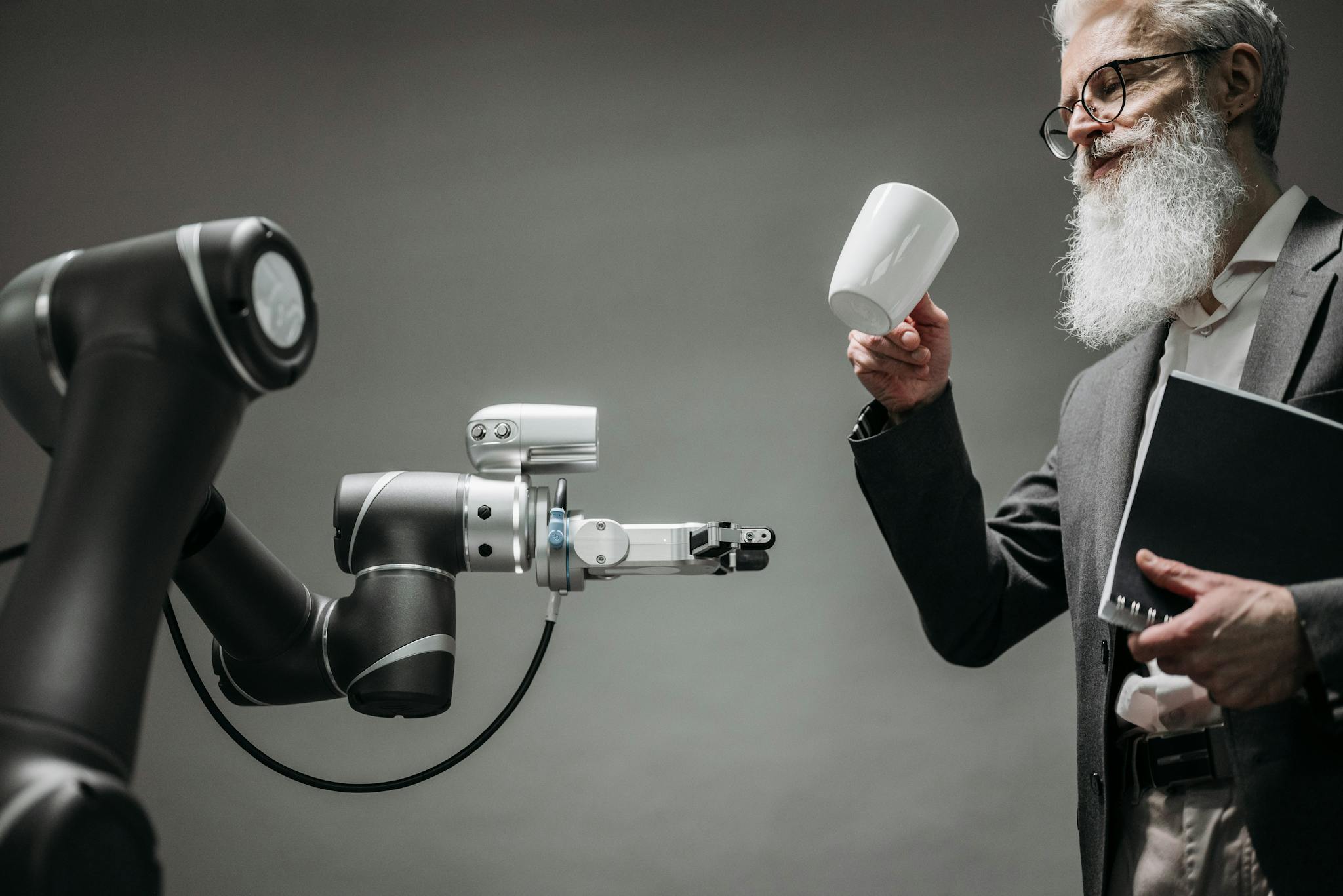 A bearded engineer holds a ceramic cup while a robotic arm assists, showcasing modern technology.